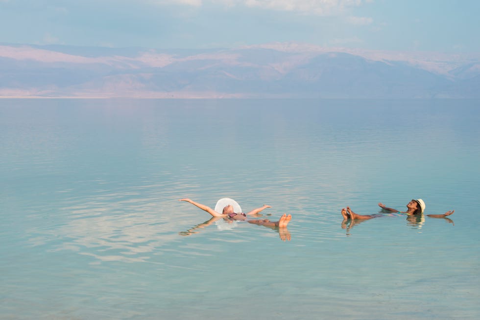 lovely multiracial girls wearing sun hats, lying on back with outstretched arms, floating in salty water of dead sea unusual buoyancy caused by high salinity