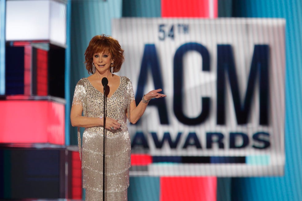 reba mcentire stands and speaks into a microphone on a stand while on a large stage, she wears a silver glittering dress and holds one hand out to the side