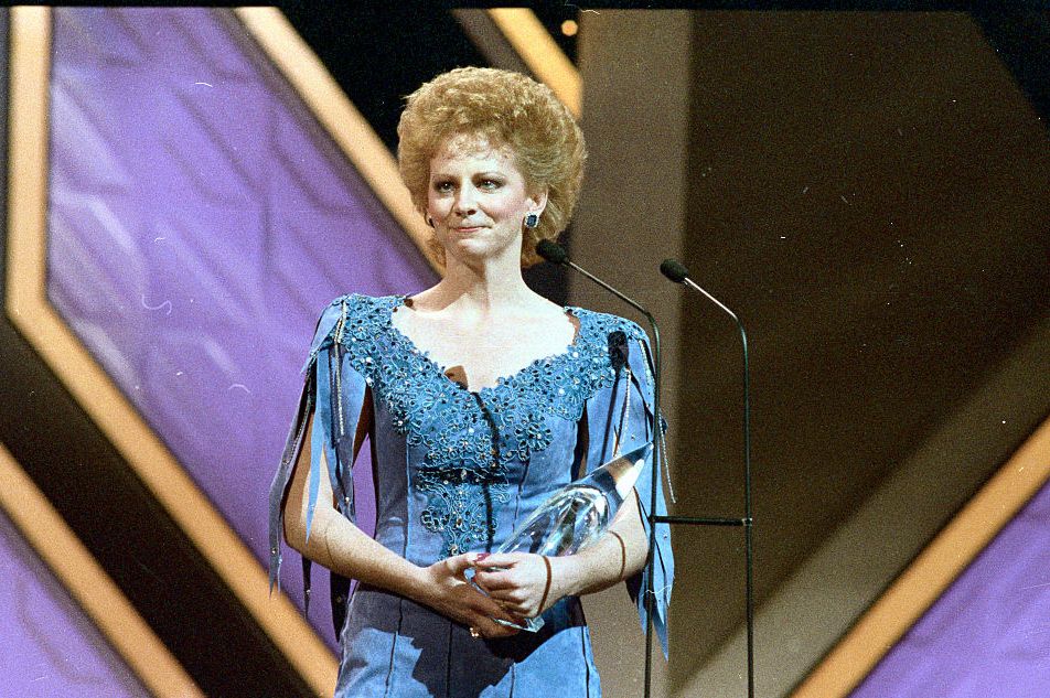 reba mcintire crying while standing on a stage, she holds a crystal award in her arms and stands behind a microphone on a stand, she wears a blue dress
