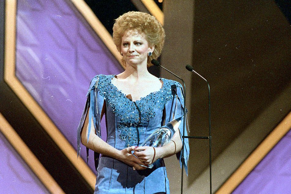reba mcintire crying while standing on a stage, she holds a crystal award in her arms and stands behind a microphone on a stand, she wears a blue dress
