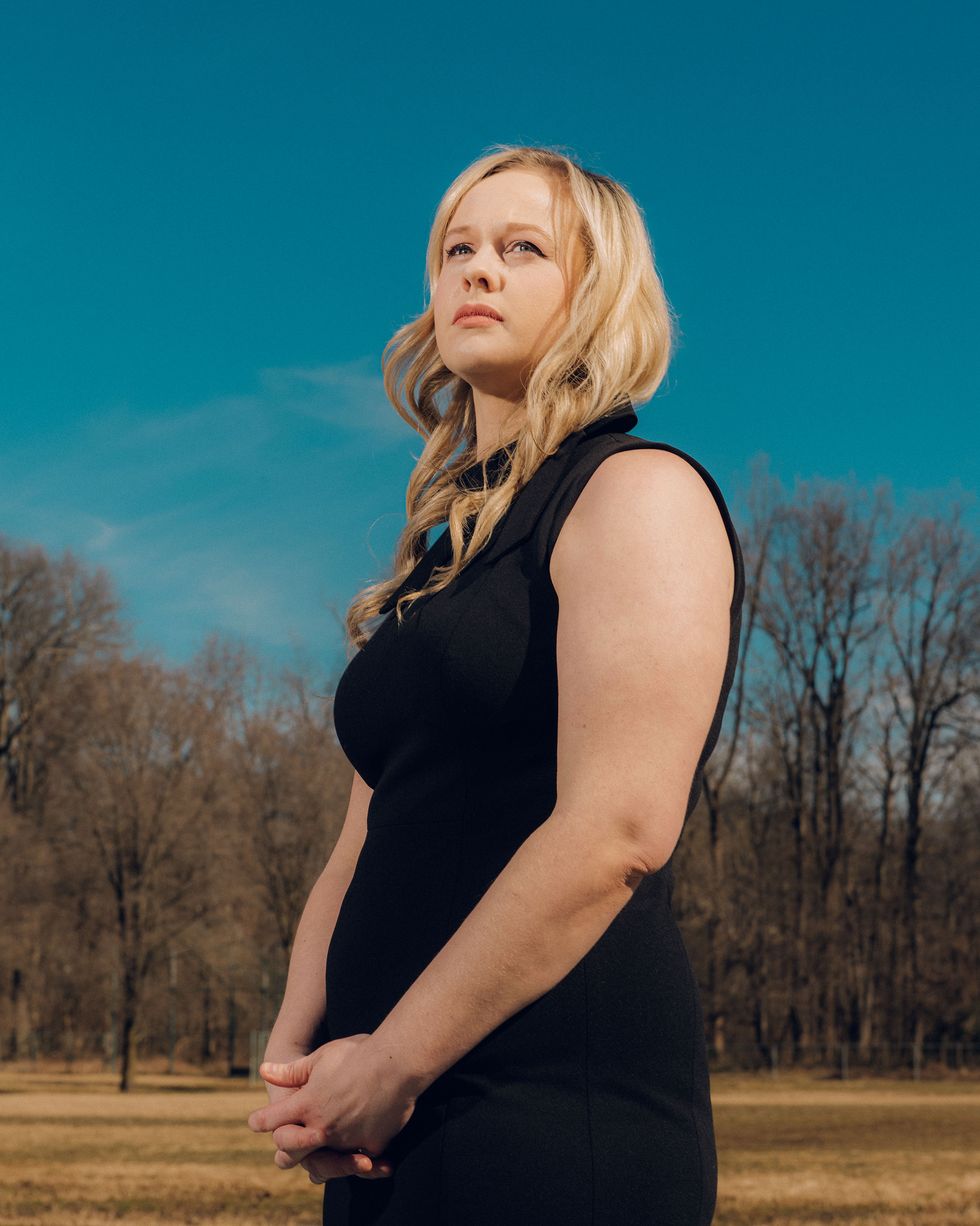 rebekah jones, unsmiling, wearing a black sleeveless dress with a small bow at the collar, standing in front of a clear blue sky and a line of trees with bare branches with her hands clasped
