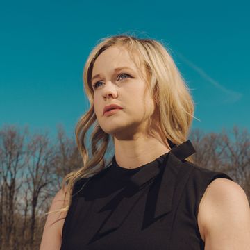 rebekah jones, unsmiling, standing in front of a clear blue sky and a line of trees with bare branches in a black sleeveless dress with a small bow at the collar