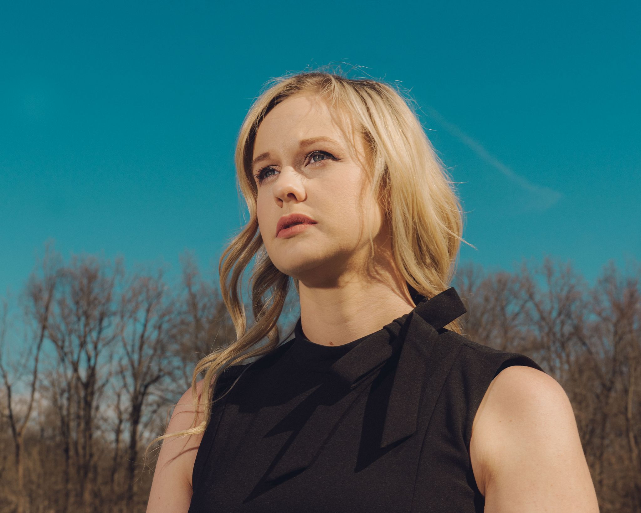 rebekah jones, unsmiling, wearing a black sleeveless dress with a small bow at the collar, standing in front of a clear blue sky and a line of trees with bare branches, looking to her right