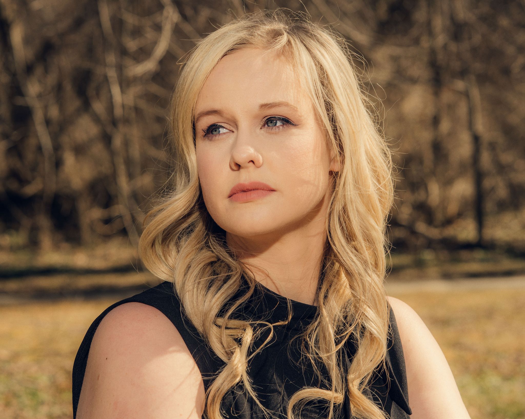 rebekah jones, unsmiling, wearing a black sleeveless dress with a small bow at the collar, sitting in front of a line of trees with bare branches