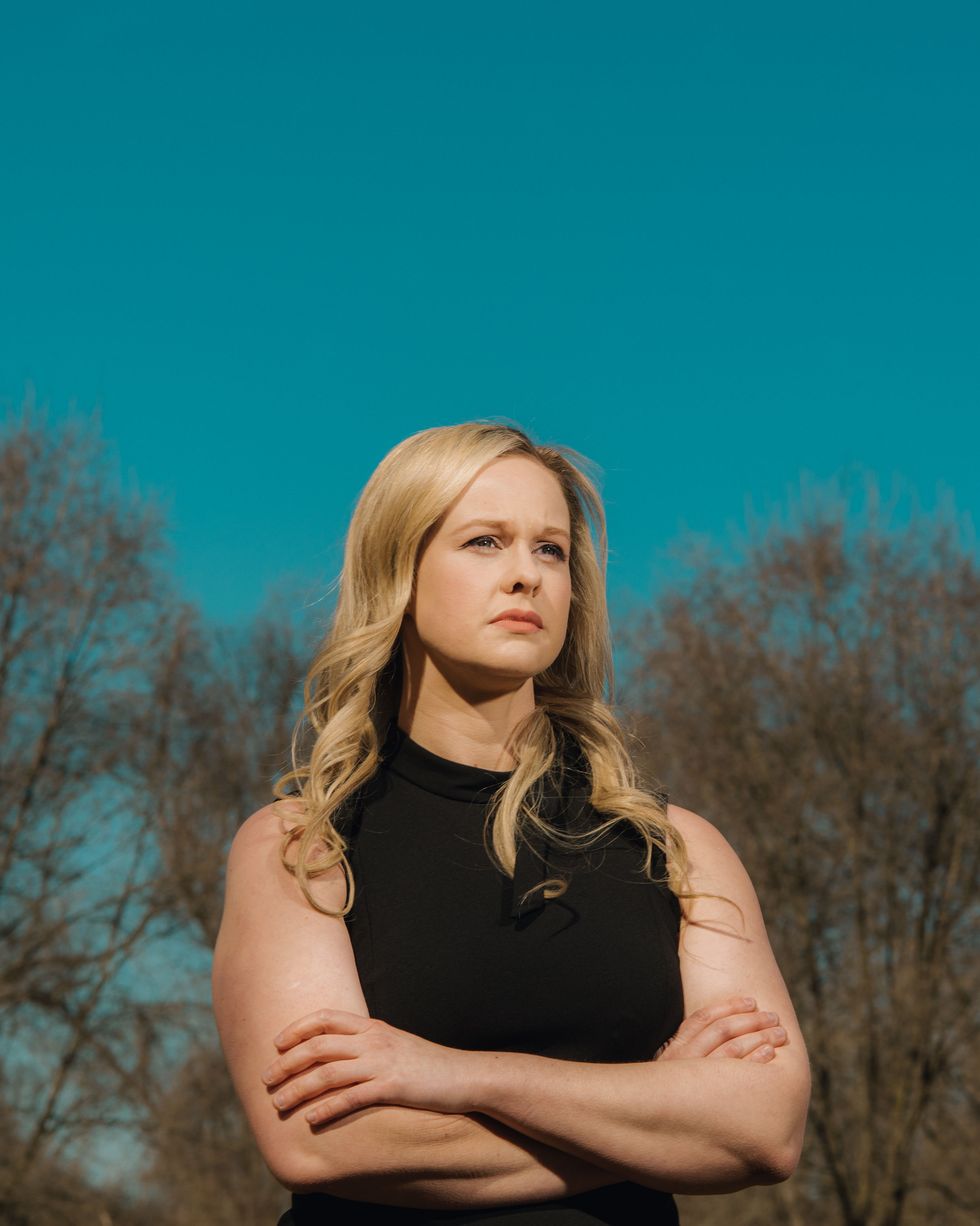 rebekah jones, unsmiling, wearing a black sleeveless dress with a small bow at the collar, standing in front of a clear blue sky and a line of trees with bare branches