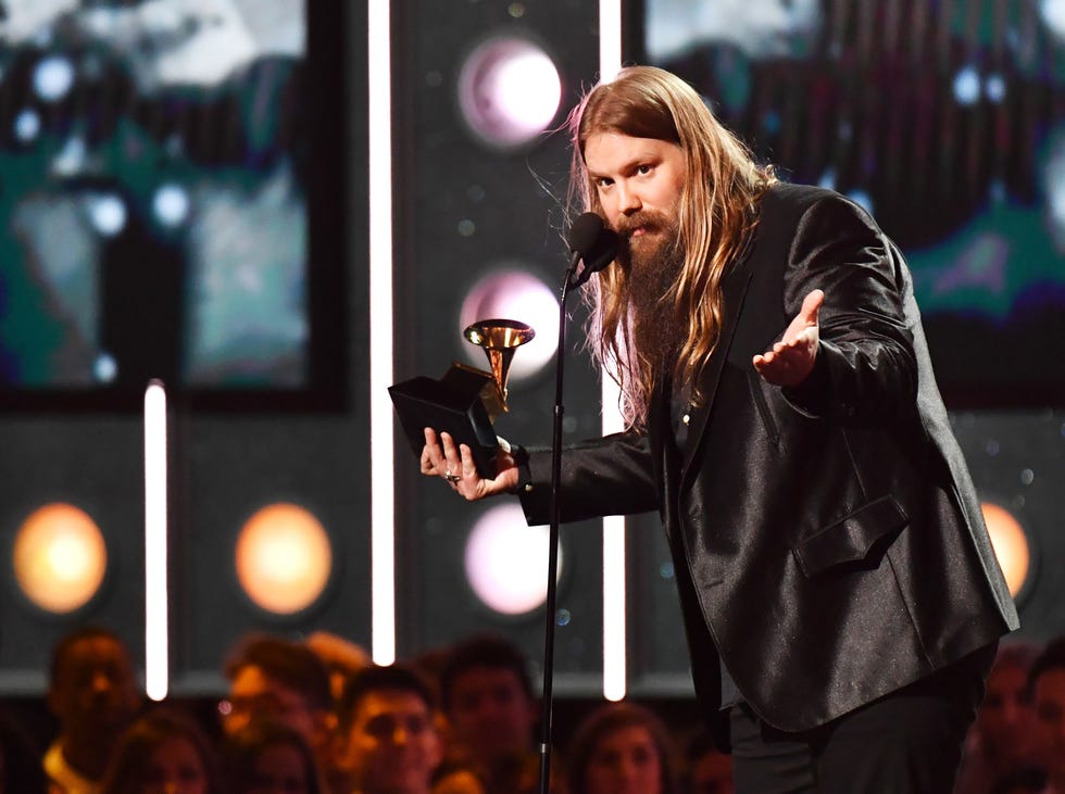 chris stapleton speaks into a microphone on a stand and gestures with his hands as he holds a grammy award in one of them