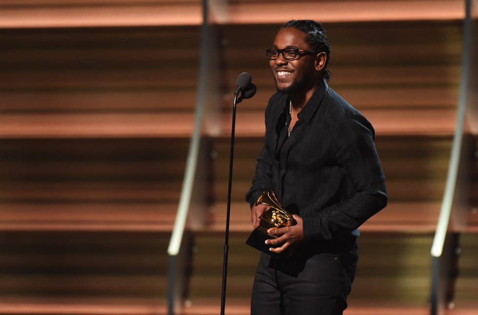 kendrick lamar holds grammy trophy and smiles while standing behind a microphone on a stand, he wears all black and dark framed glasses