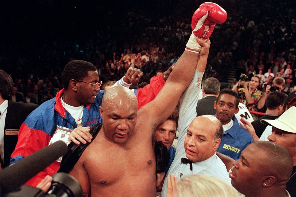 george foreman stands among a crowd of people with a robe draped on his shoulder as one fist is raised in the air with the help of another man