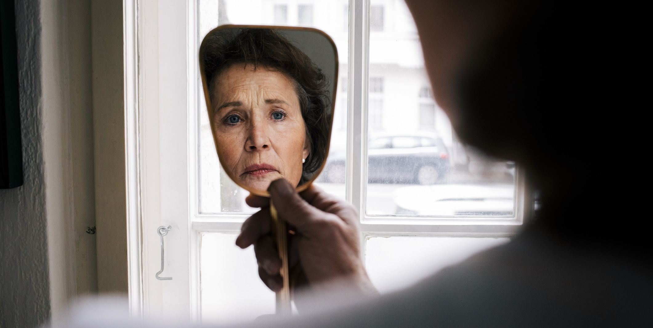 reflection of worried retired senior woman with wrinkles on hand mirror at home