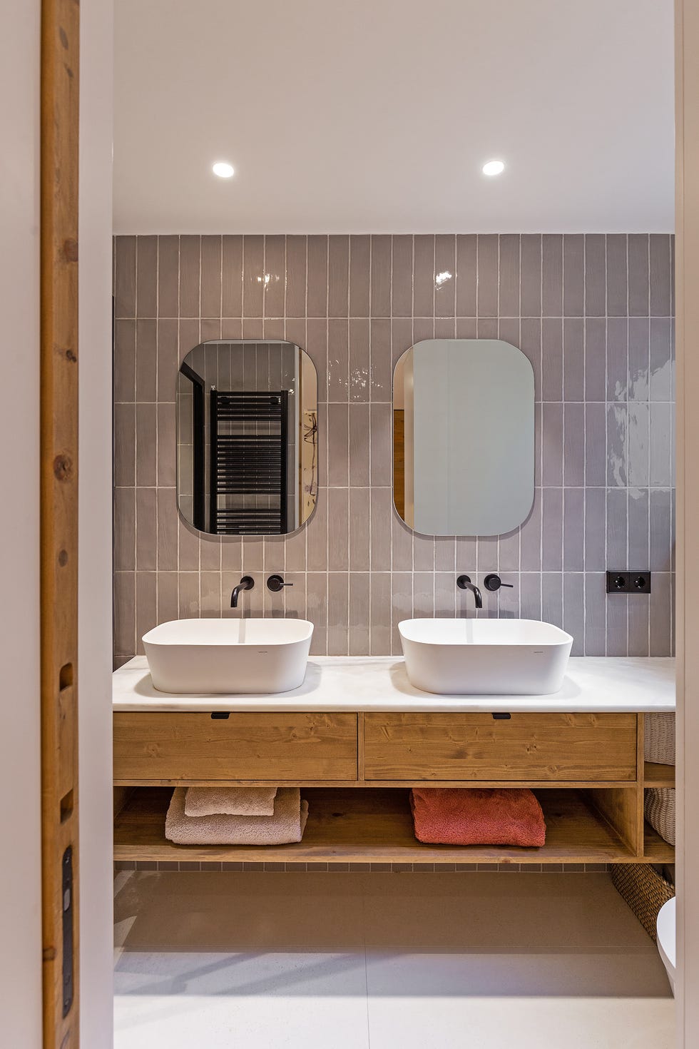baño con dos lavabos exentos, grifos negros empotrados y mueble bajolavabo de madera suspendido con azulejos grises en la pared