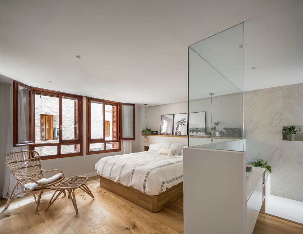dormitorio con baño en suite, suelo de madera y una butaca de fibras con reposapiés