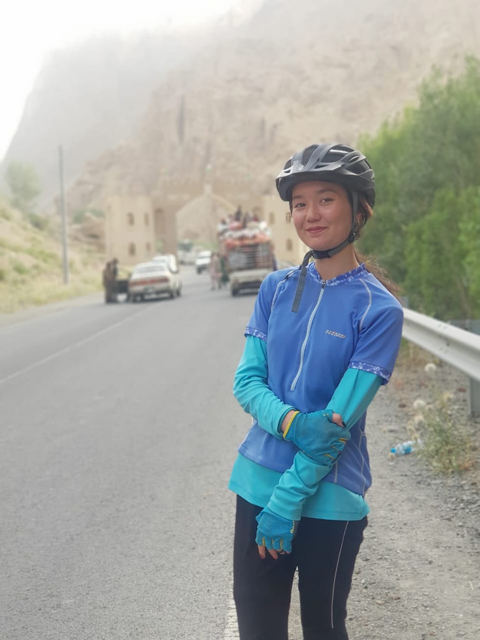 a cyclist is positioned on a roadside wearing a blue top with long sleeves and a matching helmet with a mountainous background