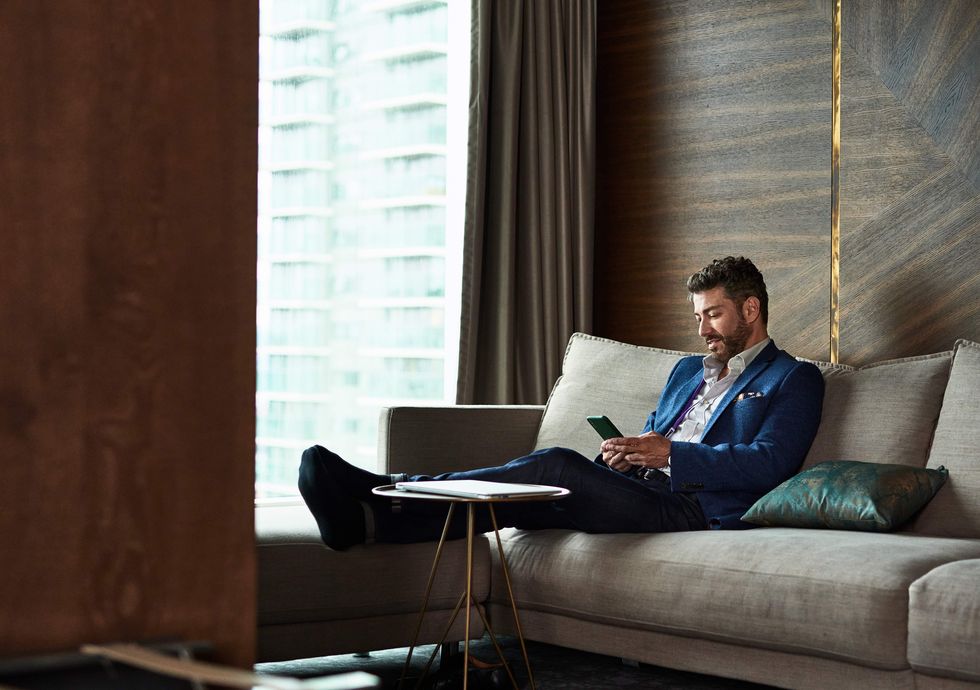 man working away from home on business trip, sitting on sofa and checking phone with feet up