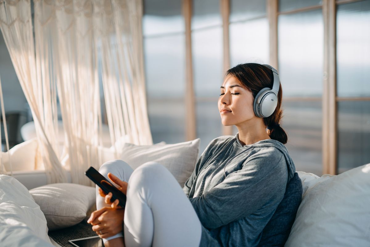 relaxed young asian woman with eyes closed sitting on her bed enjoying music over headphones from smartphone at home