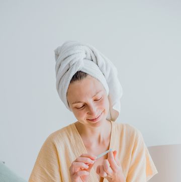 relaxing self care routine at home with woman filing nails in cozy setting. young woman in a bathrobe and a towel on her head a manicure herself and using a nail file at home.