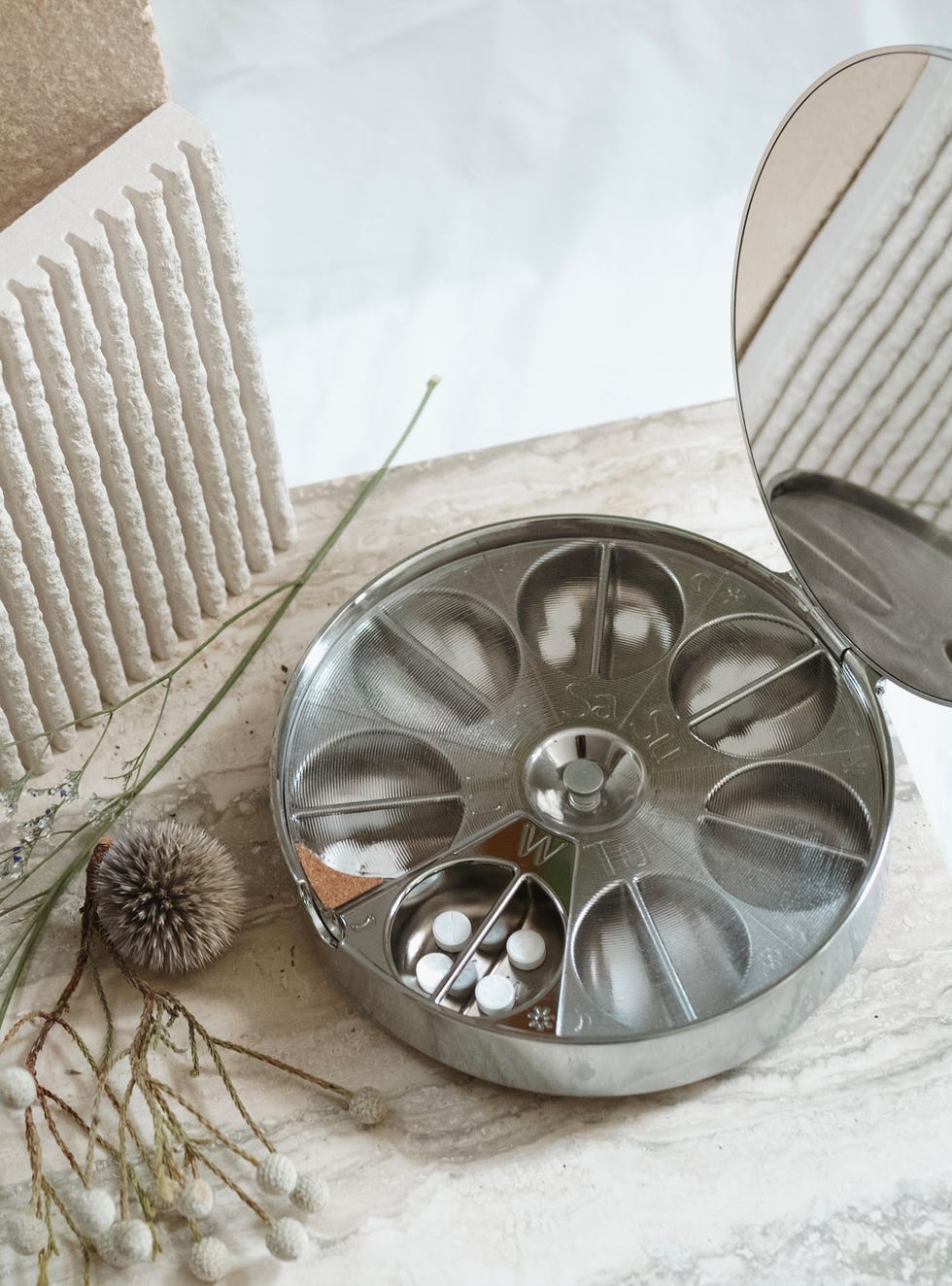 Metal pill container, with subtle weekday labels inside, a few pills in the Wednesday compartment and surrounded by dried flowers