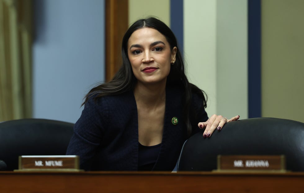 alexandria ocasio cortez preparing to sit down behind a panel with multiple chairs