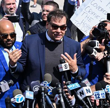 george santos, wearing a blue suit jacket, black shirt, and sunglasses, speaks in front of several microphones amid a large crowd