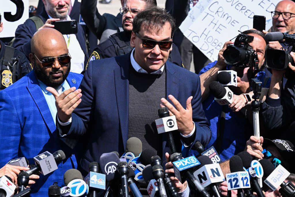 george santos, wearing a blue suit jacket, black shirt, and sunglasses, speaks in front of several microphones amid a large crowd