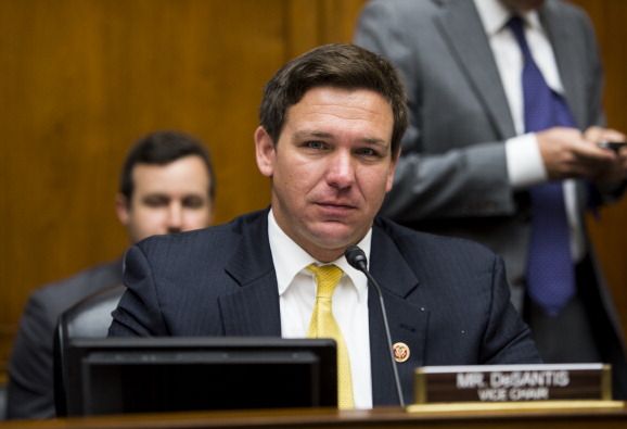 ron desantis stares at the camera while sitting in a wood paneled room, he has a display screen, microphone and name plate in front of him, he wears a black suit jacket, white collared shirt and yellow tie