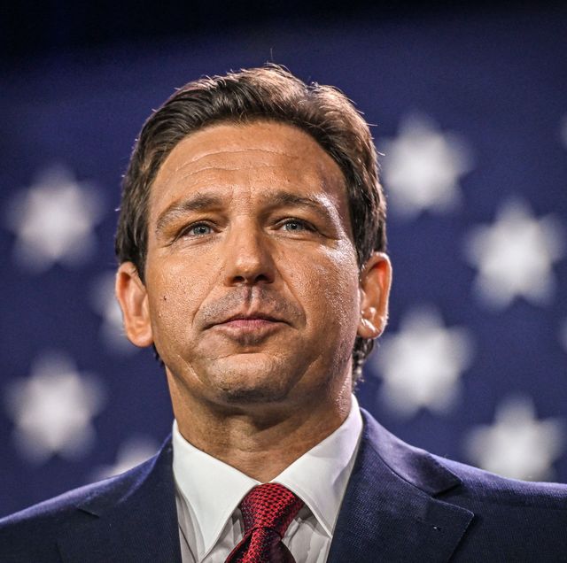ron desantis looks past the camera with a slight smile, he wears a navy suit jacket with a white collared shirt and red tie, in the background is part of a large american flag that is out of focus