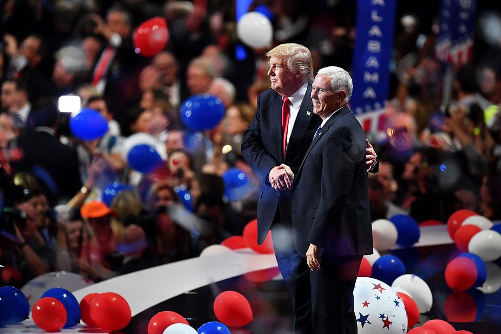 a profile shot of donald trump and mike pence standing on a stage shaking hands, both men wear suits, on the stage floor around them are red, white, and blue balloons and behind them is a blurred crowd