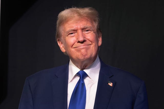 donald trump smiles at the camera, he wears a blue suit jacket and tie with a white dress shirt, an american flag pin is on his lapel