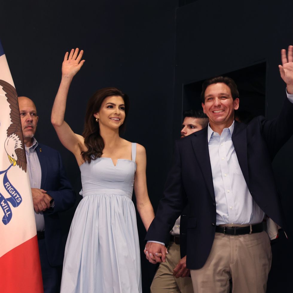 casey desantis and ron desantis hold hands, smile, and wave to a crowd out of view, she wears a baby blue dress and he wears a suit jacket with a light blue collared shirt and khaki pants