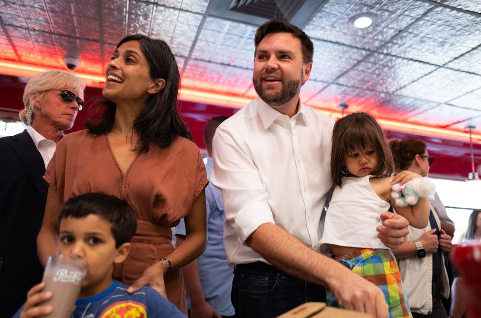 Usha Vance and JD Vance smile while carrying two of their young children with them in a crowded restaurant