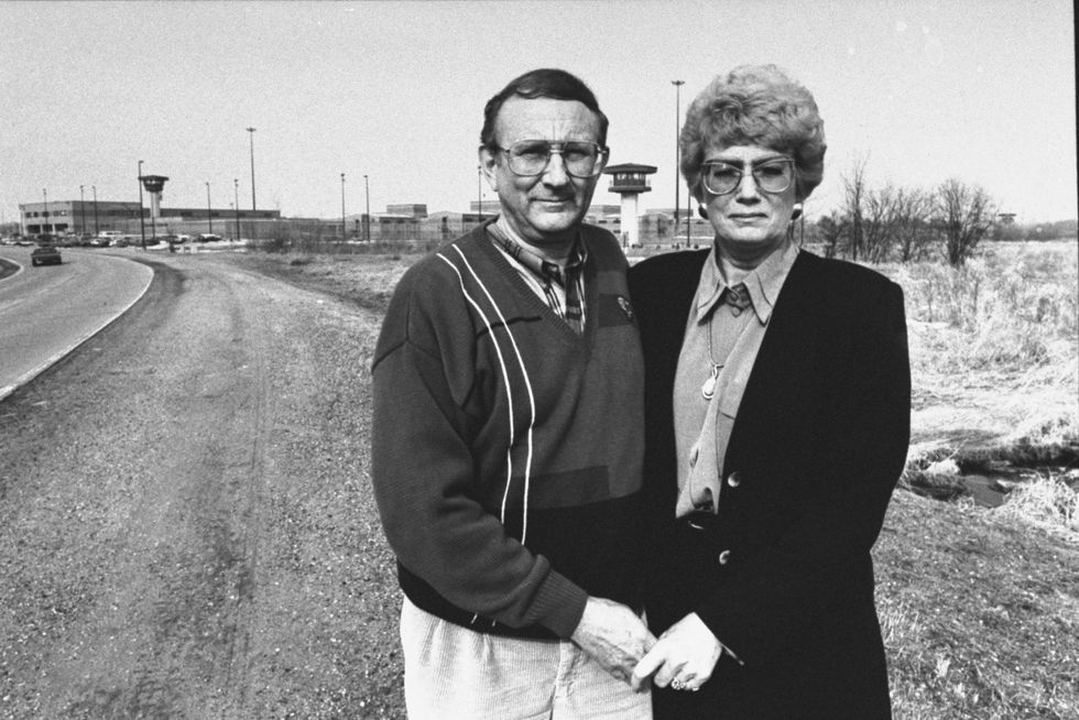 jeffrey dahmer's father and stepmother stand outside in front of a prison, they are hugging each other and holding hands while looking at the camera
