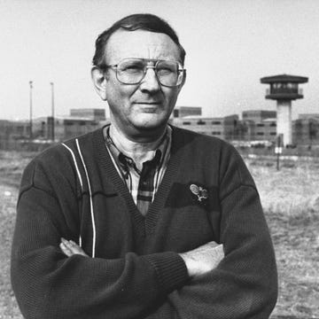 lionel dahmer stands outside a fenced complex with his arms crossed, he wears a sweater, plaid shirt, and large glasses