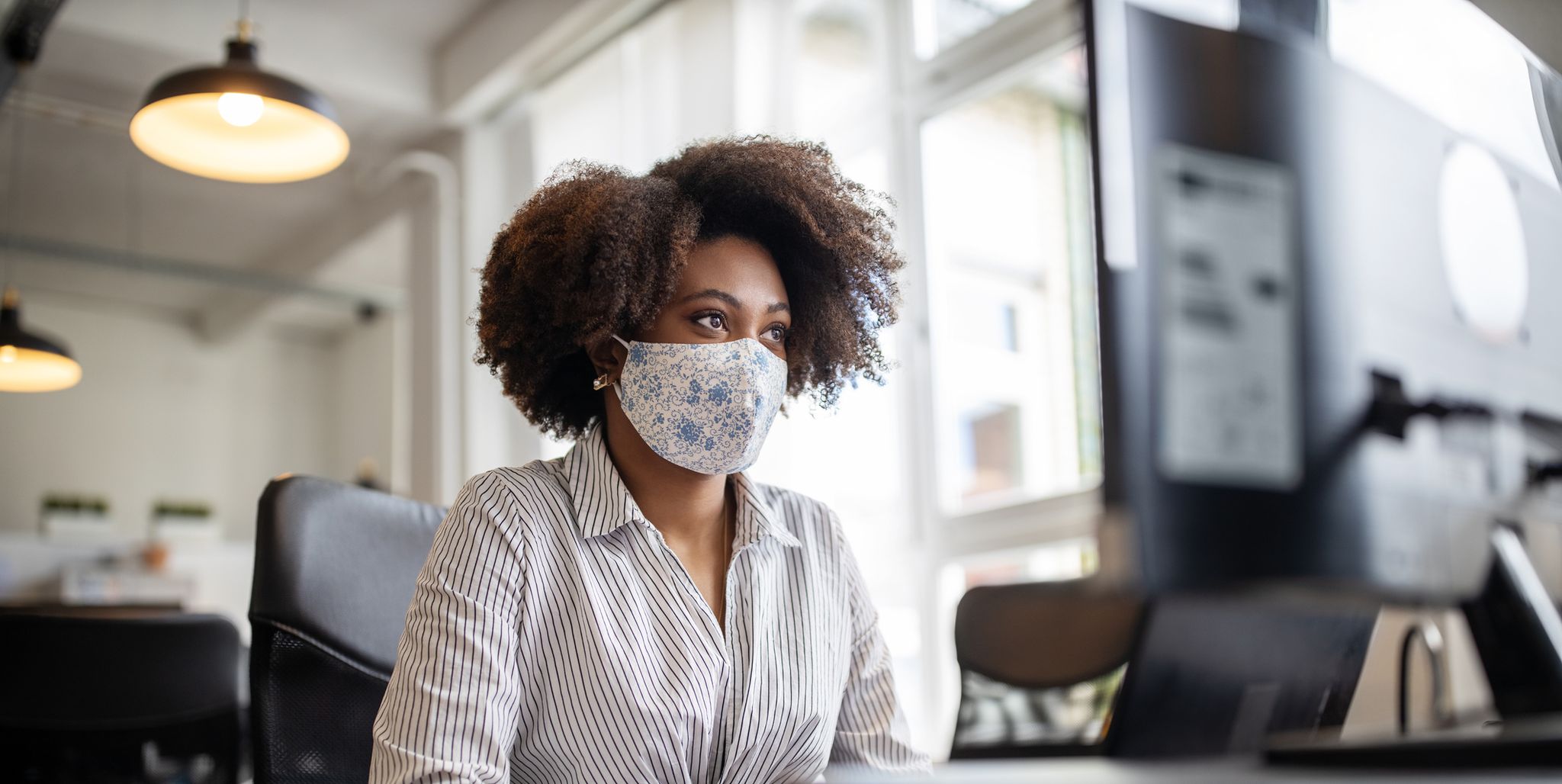businesswoman with face mask working at her desk looking at computer monitor, in office female professional back to work after covid 19 pandemic lockdown