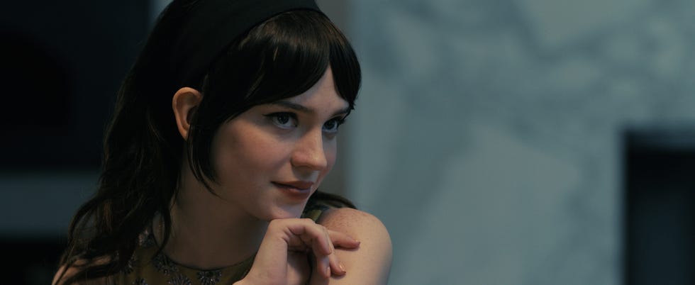 A person with dark wavy hair and a headband seated possibly in a contemplative state