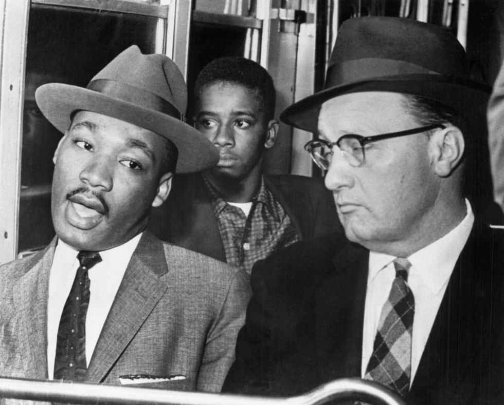 martin luther king jr talks to a white man sitting next to him on a bus, both men wear suits with ties and hats, a man sits behind them and looks out the window
