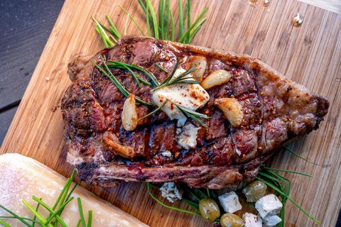 ribeye steak just off a charcoal grill resting on a wood cutting board, seasoned with rosemary, garlic cloves, butter, cheese, olives and paprika