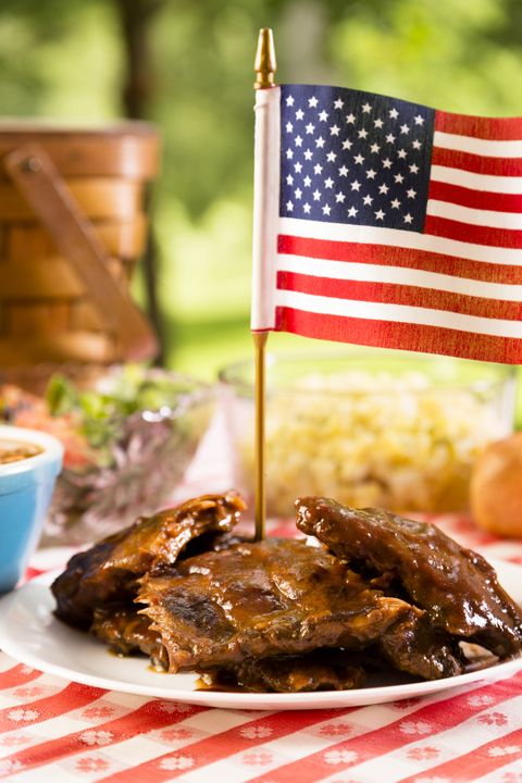 bbq ribs topped by american flag, beans, potato salad on a red and white table cloth