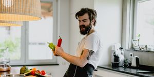 handsome bearded young man preparing healthy meal with vegetables, using headphones listening to music and singing in the kitchen at home
