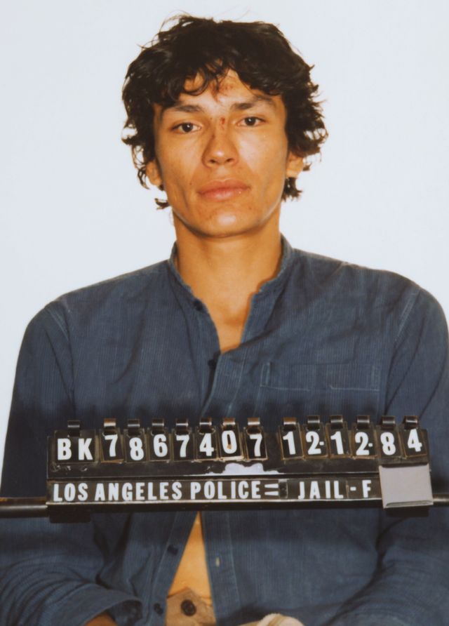 richard ramirez stares at the camera in a police mug shot, he is wearing a blue corduroy button up shirt and his inmate number is in front of his chest on a suspended bar