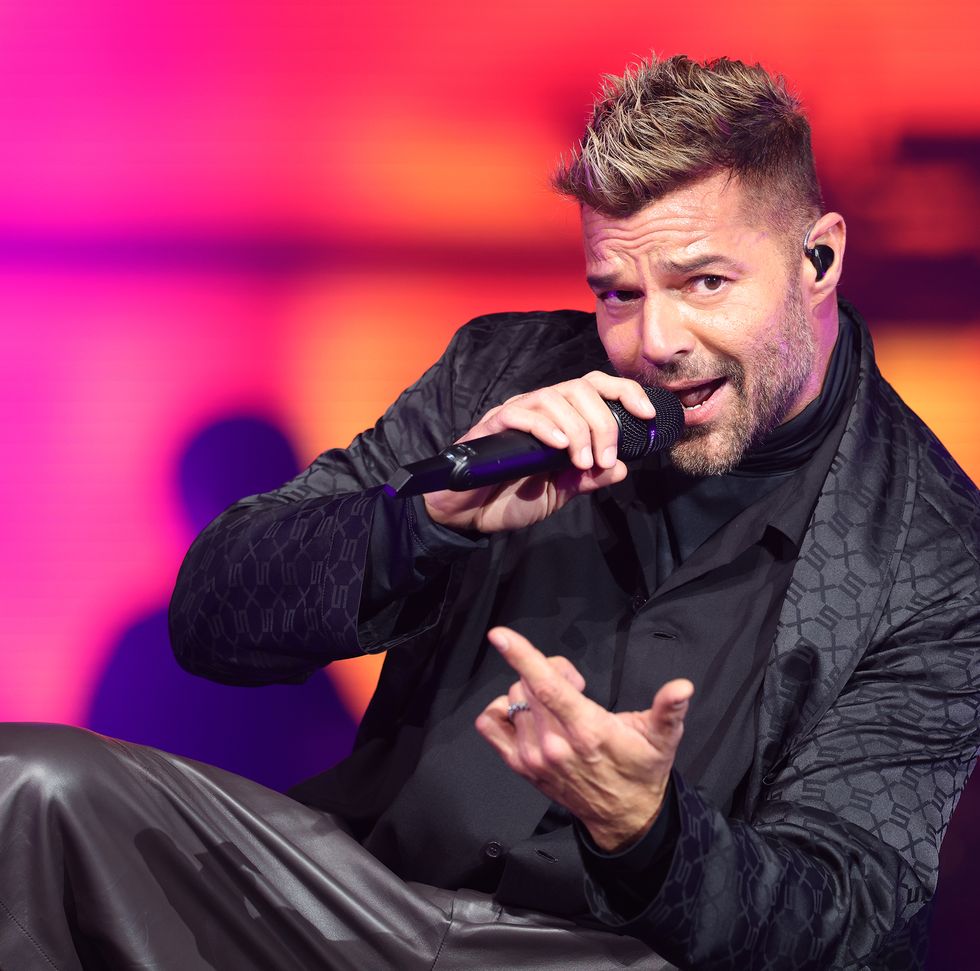 a man in all black sings into a wireless microphone as he crouches down while on a pink and orange lit stage