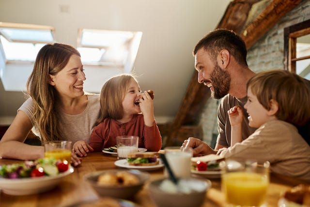 young parents and their two small kids, a girl and a boy, talking and laughing while having meal at table