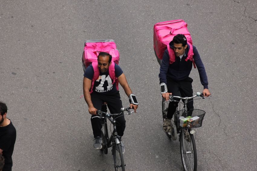 Foto con due fattorini in bicicletta in una strada di Milano indossano due enormi zaini cubici che contengono il cibo da consegnare a domicilio. 
