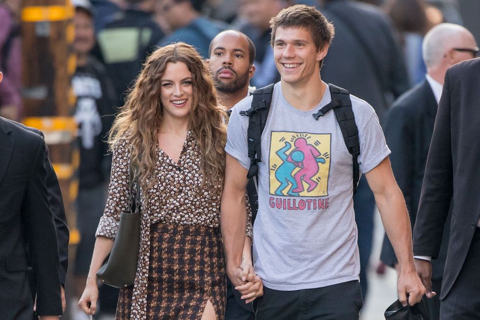 riley keough and ben smith petersen smile and walk hand in hand on a sidewalk, she wears a patterned button up shirt with a houndstooth pencil skirt, he wears a gray graphic t shirt and a backpack
