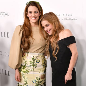 riley keough and lisa marie presley smile and hug for photos, they stand in front of a white background with black written logos, keough wears a tan long sleeve blouse with a green and yellow floral skirt, presley wears a back off the shoulder dress
