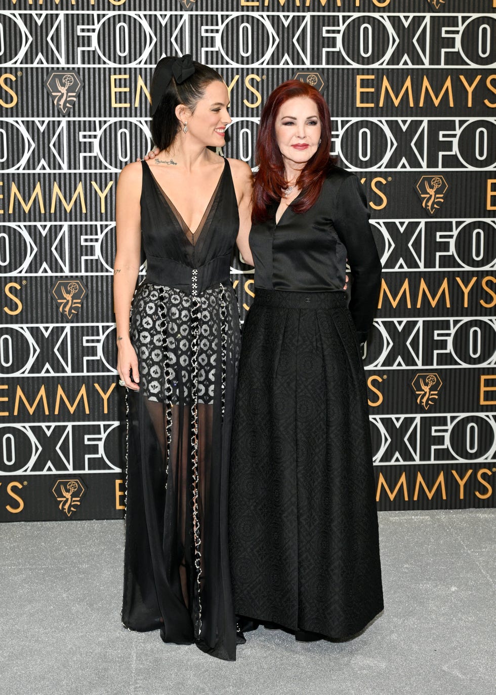 riley keough smiles and looks right at priscilla presley who smiles for photos, both women embrace and wear formal black dresses while standing in front of a black background with with and gold writing
