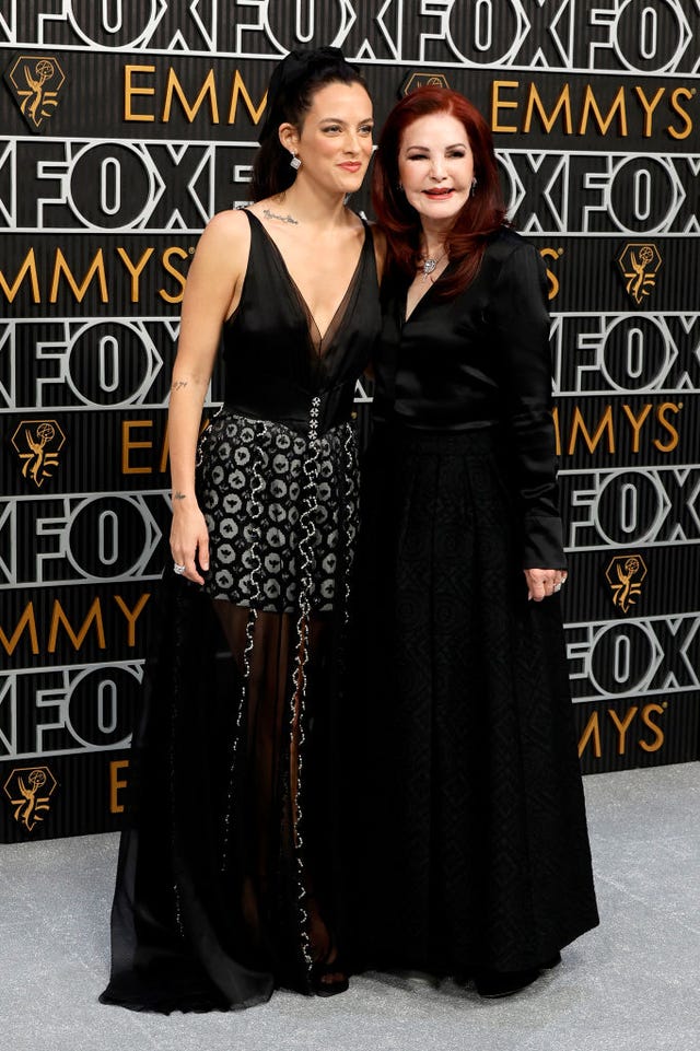 riley keough and priscilla presley smile for photos while standing in front of a black, white and gold photo background, both women wear formal black dresses and hug one another