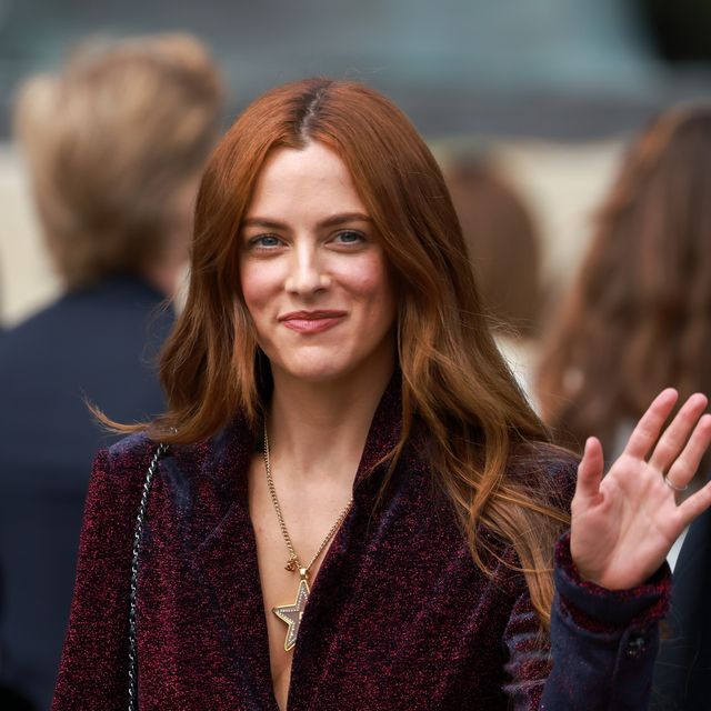 riley keough smiles and waves at the camera, she wears a burgundy jacket and golden star necklace