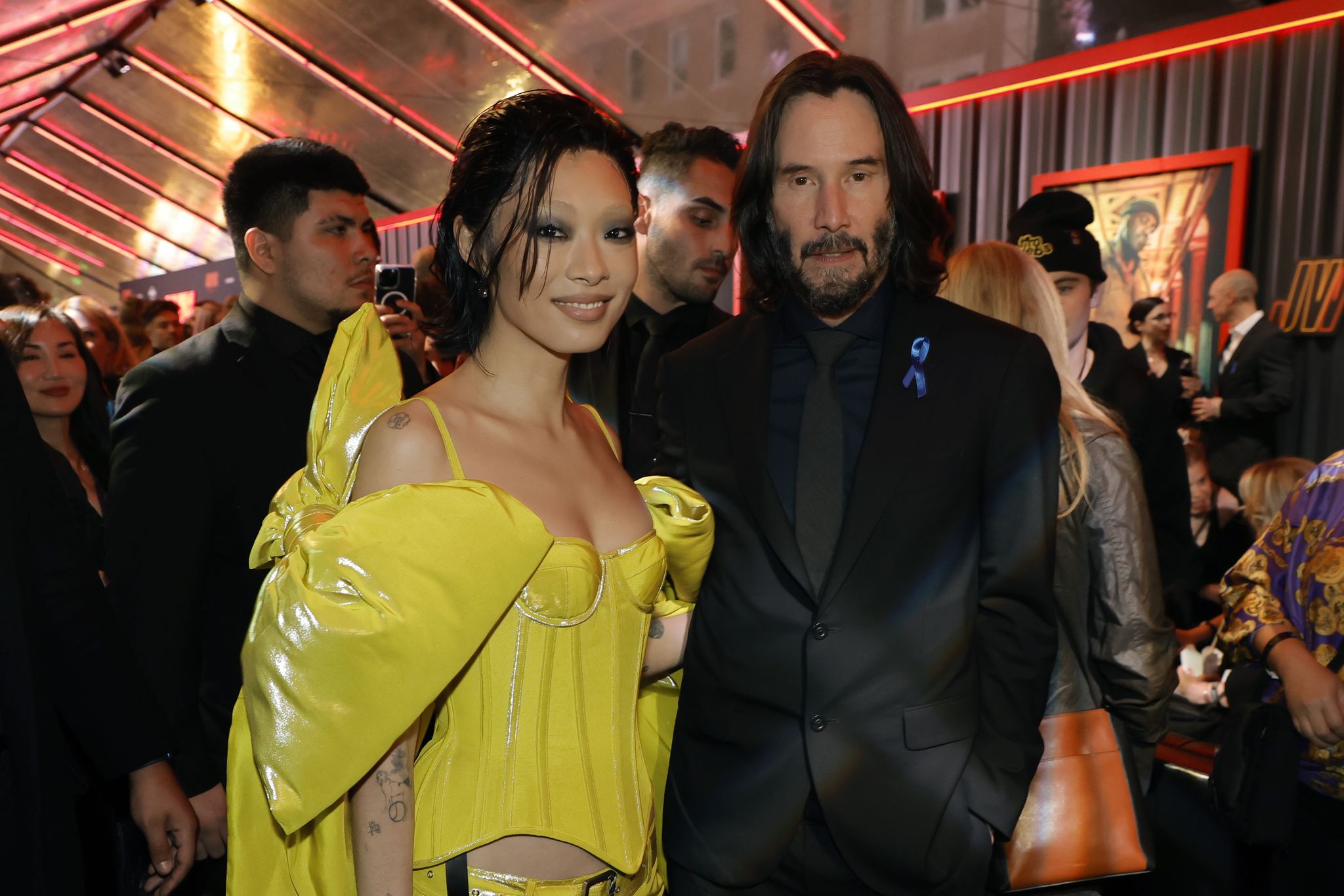 rina sawayama wearing a yellow dress, and keanu reeves wearing a black suit and tie, in a crowded movie theater