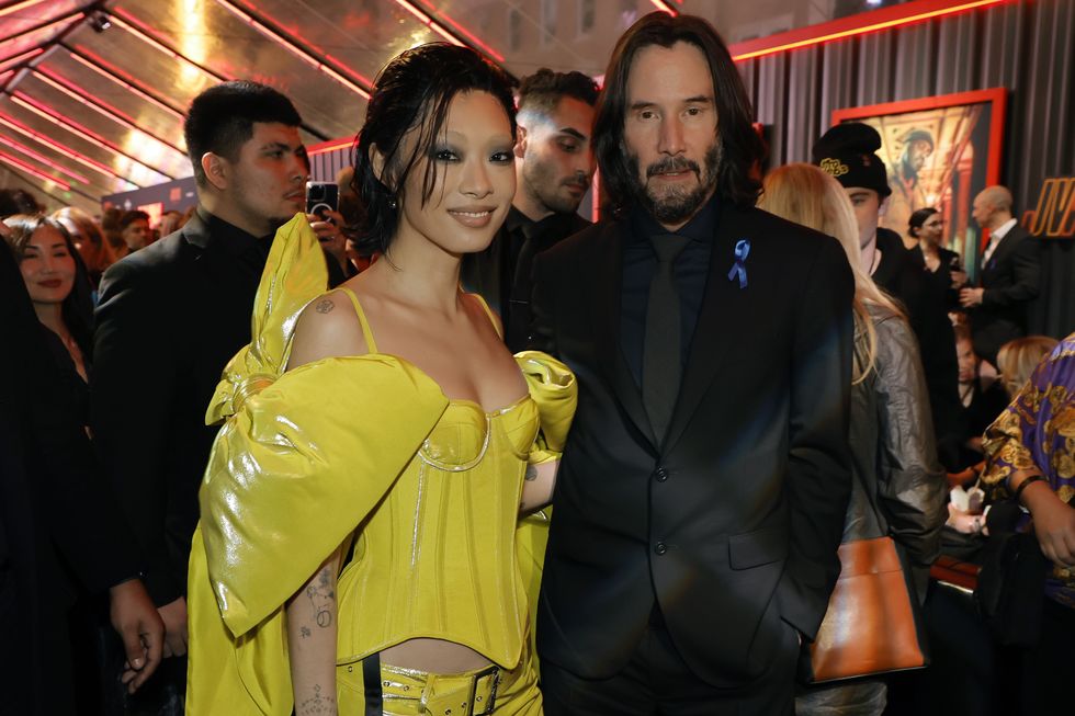 rina sawayama wearing a yellow dress, and keanu reeves wearing a black suit and tie, in a crowded movie theater