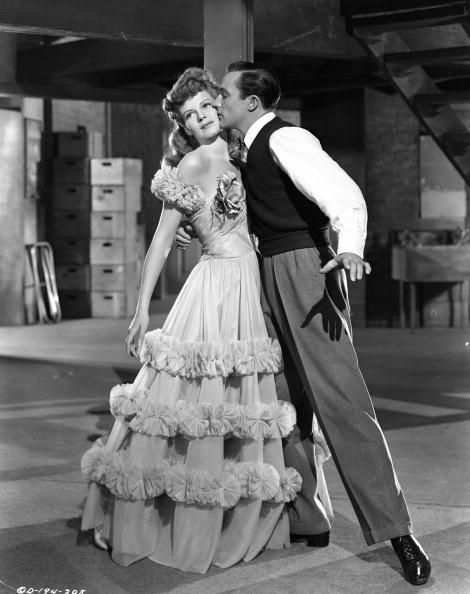 rita hayworth stands next to gene kelly who wraps one arm around her waist and kisses her cheek, she wears a ball gown, he wears slacks, a white collared shirt, and a dark sleeveless sweater vest