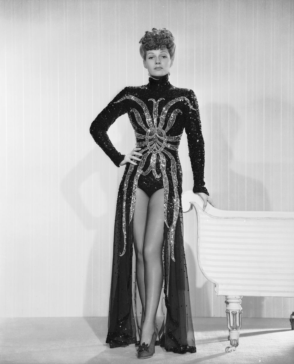 rita hayworth stands next to a piece of furniture that she holds with one hand, her other hand is on her hip, she wears a sequined outfit with a spider design on the front